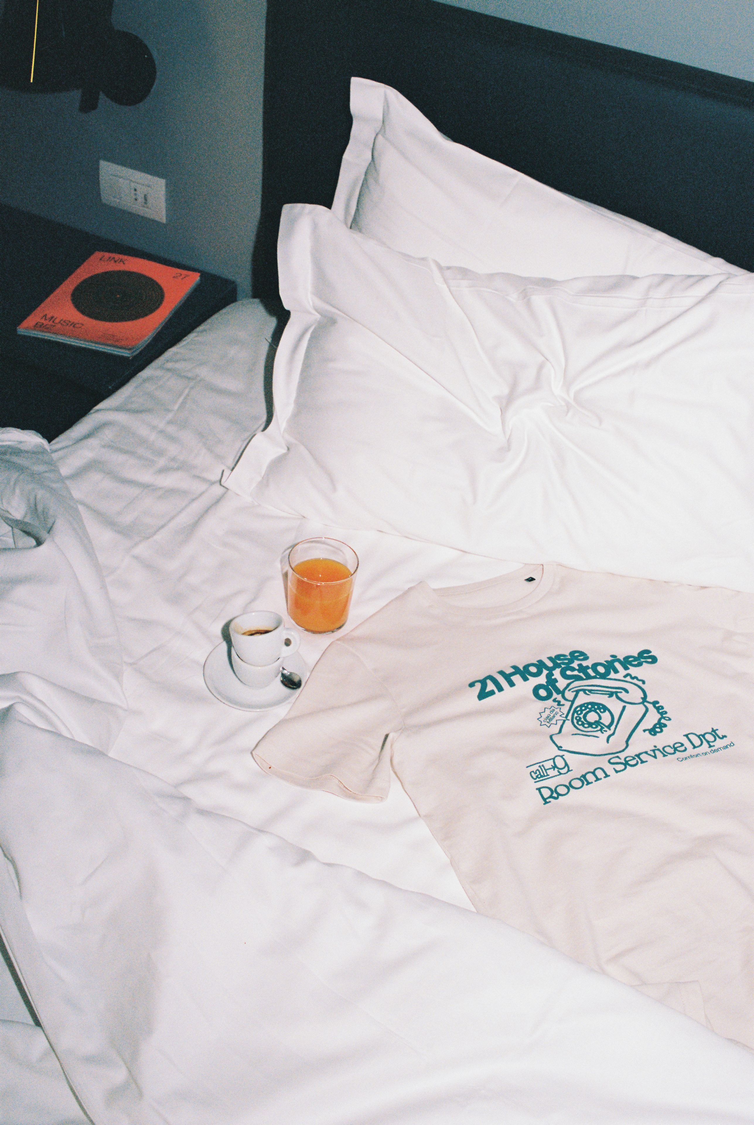 A bed with white linens, a glass of orange juice, a cup of coffee, and a t-shirt with a logo.