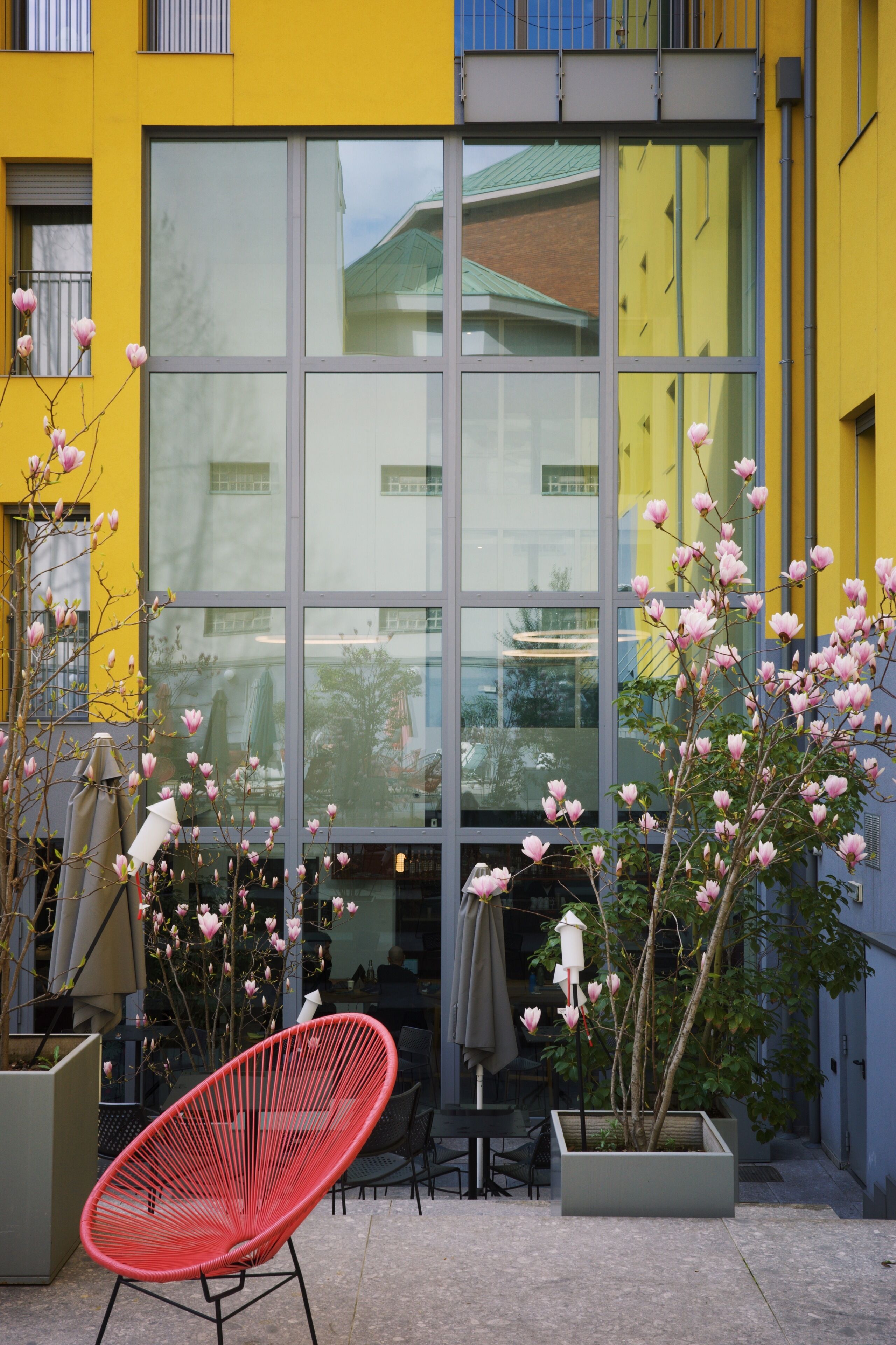 A yellow building with large windows, a red chair, and a potted plant with pink flowers.