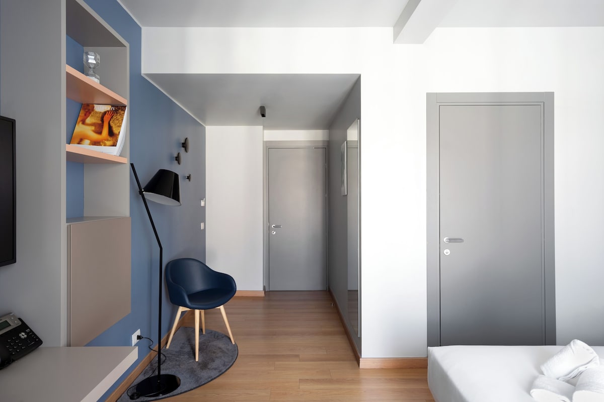 Un dormitorio moderno con cama, escritorio con computadora, silla, armario y un pasillo que lleva a otra habitación.