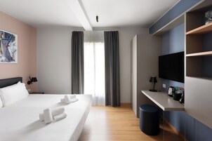 Et hotelværelse med seng, skrivebord, fjernsyn og et vindue med gardiner.