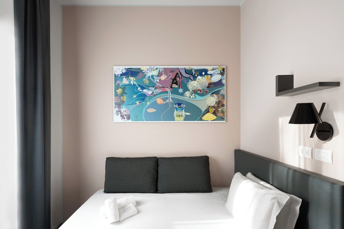 Habitación de hotel con cama, almohadas, una obra de arte en la pared, lámpara y estante.