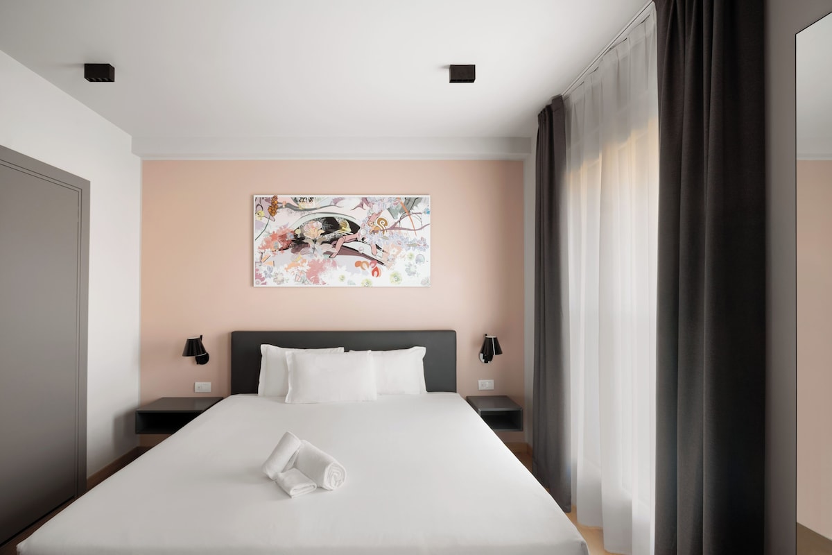 Una habitación de hotel con cama, una obra de arte en la pared, una mesita de noche y una ventana con cortinas.
