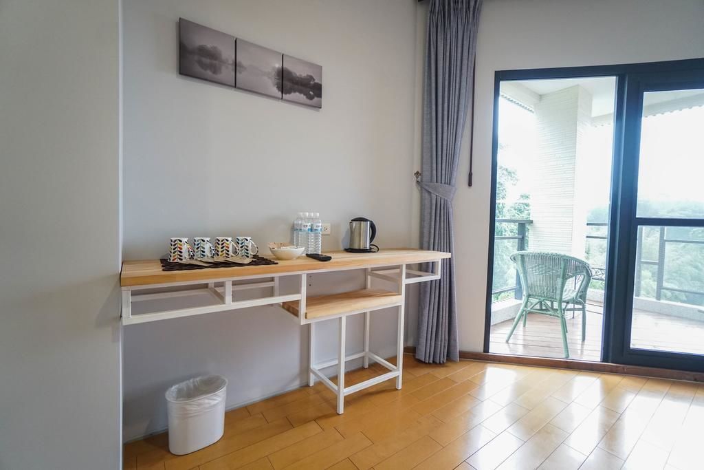 A room with a wooden desk, a white trash bin, and a view of a balcony with a chair.