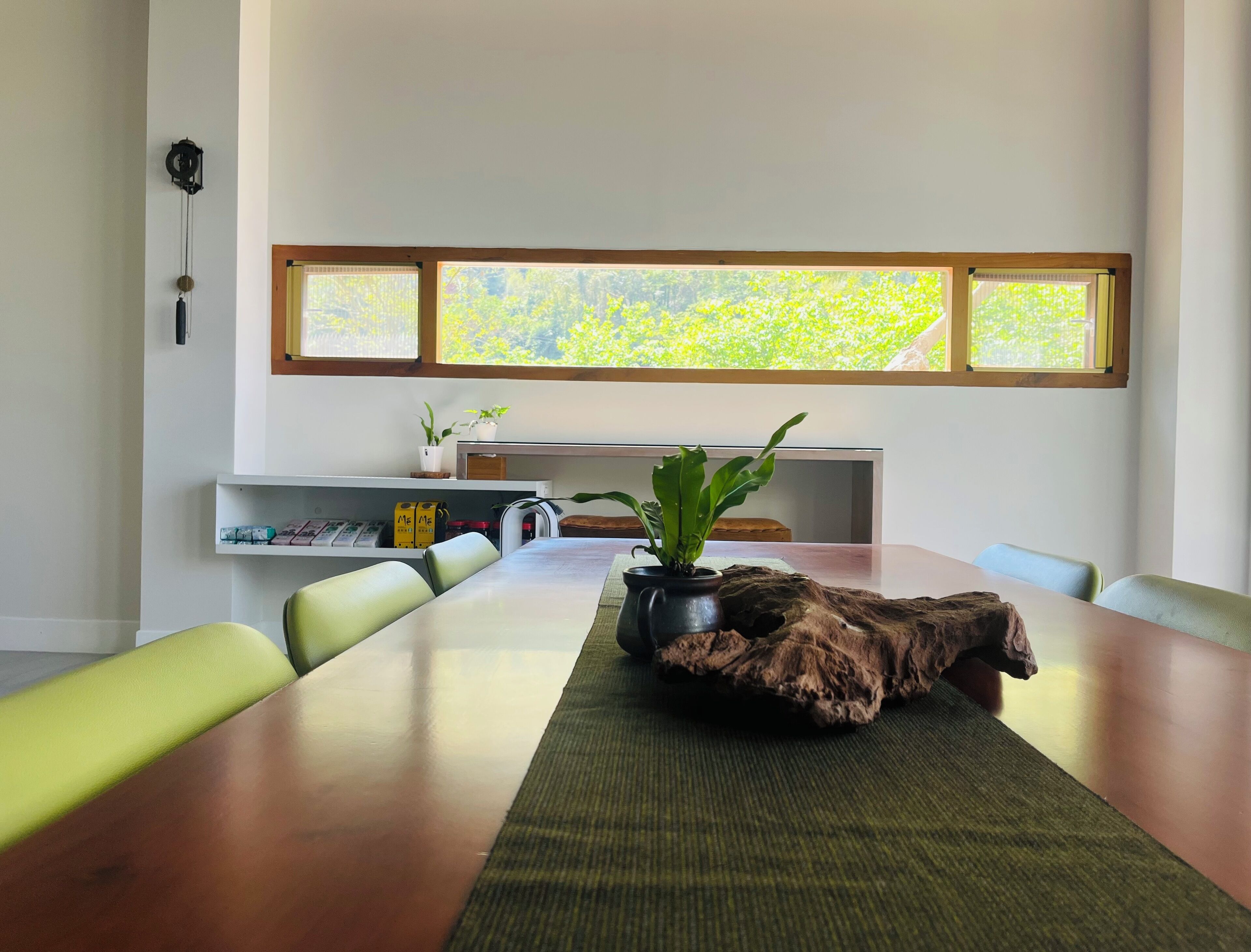 A dining area with a long table, green chairs, a potted plant, a wooden table runner, and a view of greenery through windows.