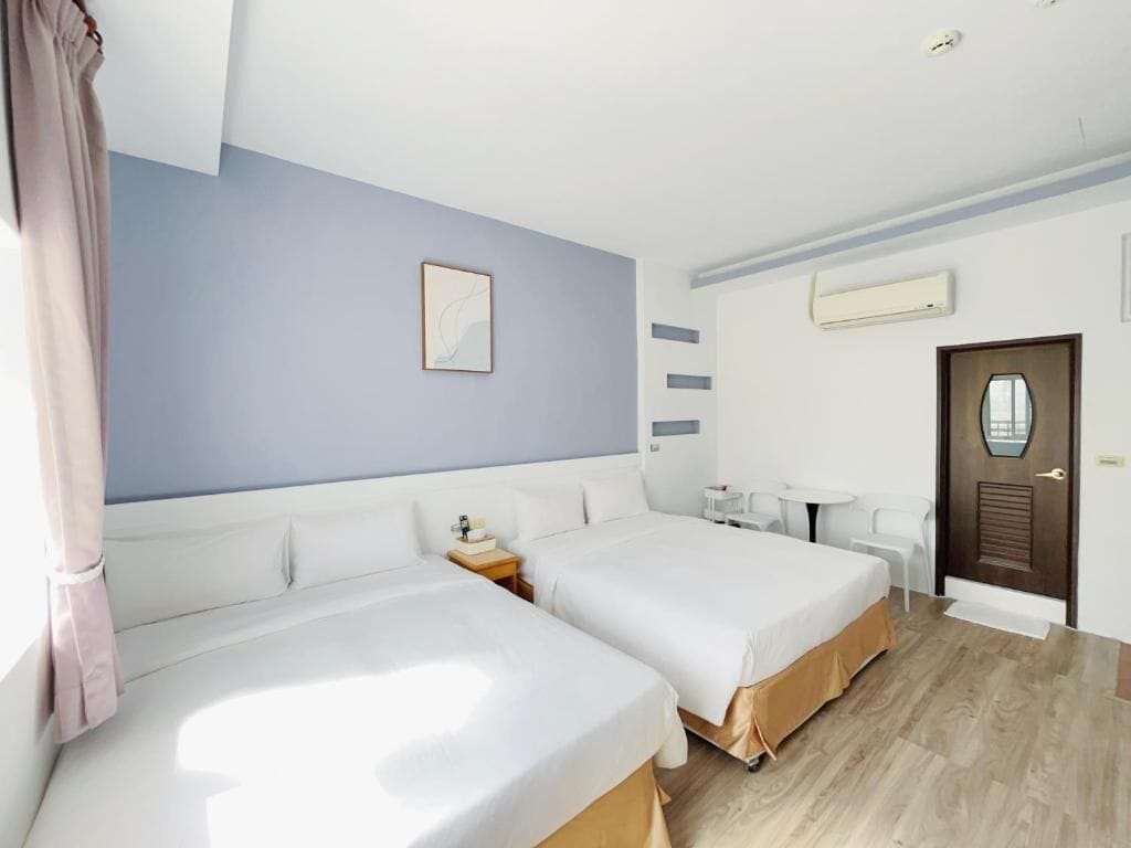 Quarto de hotel com duas camas, uma escrivaninha, uma cadeira, uma mesinha e uma porta.