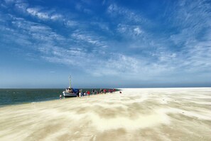 Ein Strand mit einem angestauten Boot auf der linken Seite, Menschen, die am Sand entlanggehen, und einem wolkenlosen, blauen Himmel.