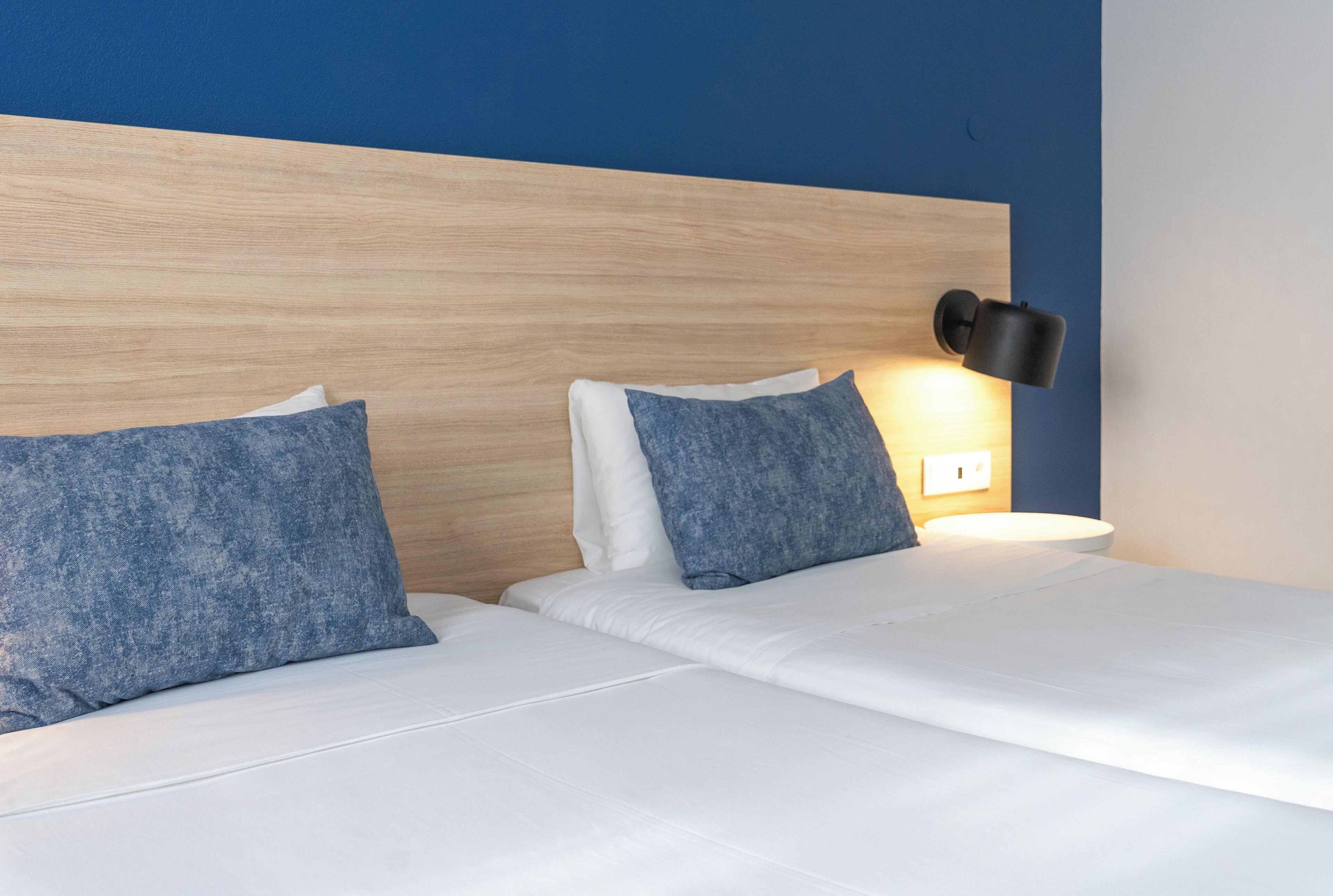A hotel room with a wooden headboard, two pillows, a bedside lamp, and a white bedspread.