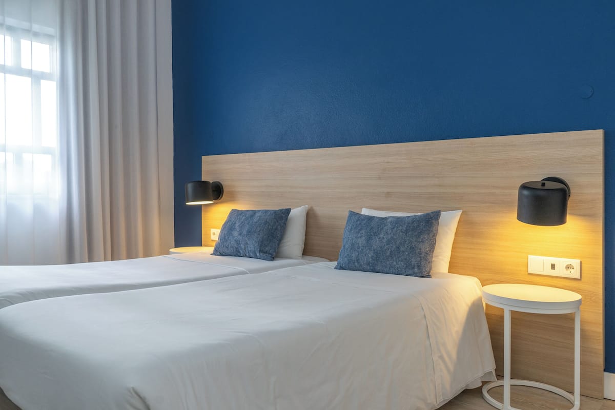 Hotellihuone, jossa on kaksi sänkyä, yöpöytä, seinään kiinnitetty valaisin ja pistorasia.