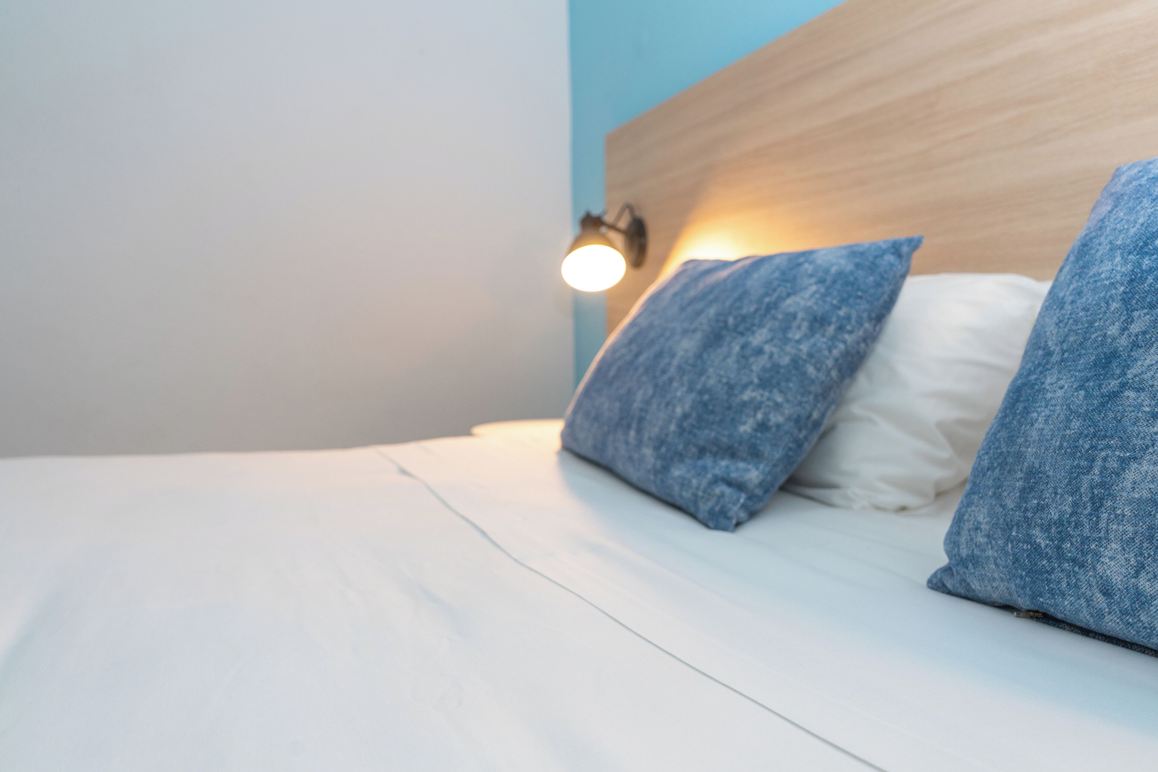 Un letto rifatto con lenzuola bianche e due cuscini blu, una lampada a parete e una testiera in legno.