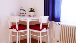A small, white table with two white chairs, each with a red cushion. A white pot with pink flowers and a white candle are on the table. A blue curtain partially covers a window. - Adventure Guesthouse (Unterseen)