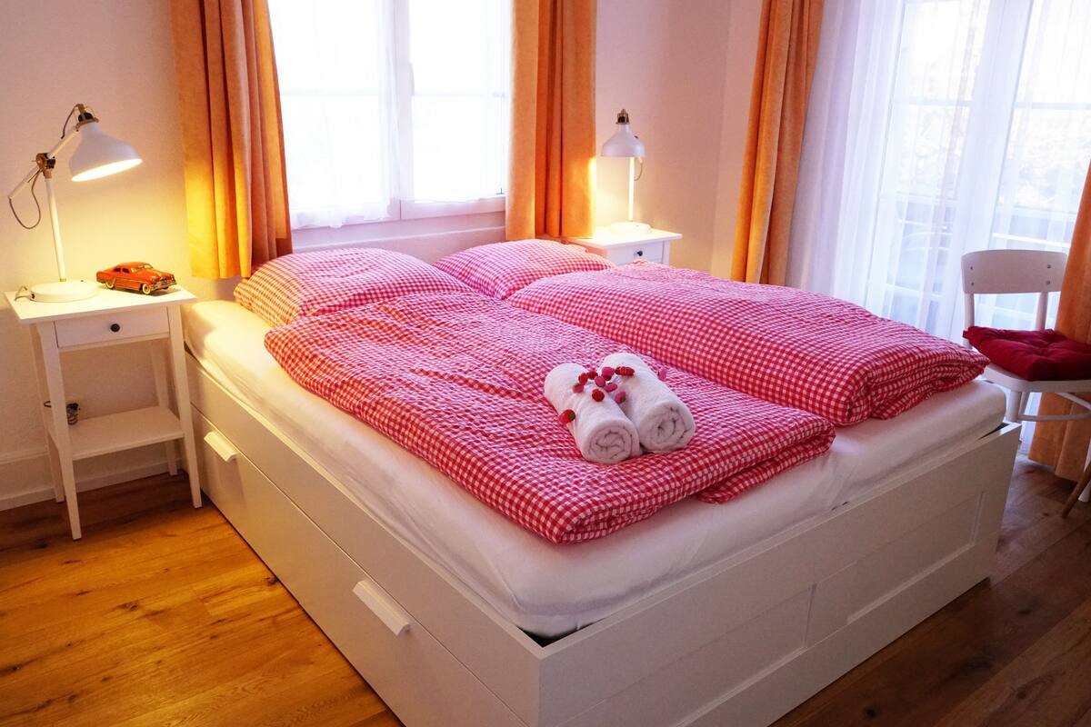 Uma cama bem arrumada com roupa de cama xadrez vermelha, duas luminárias de cabeceira e uma mesinha com um carrinho de brinquedo.