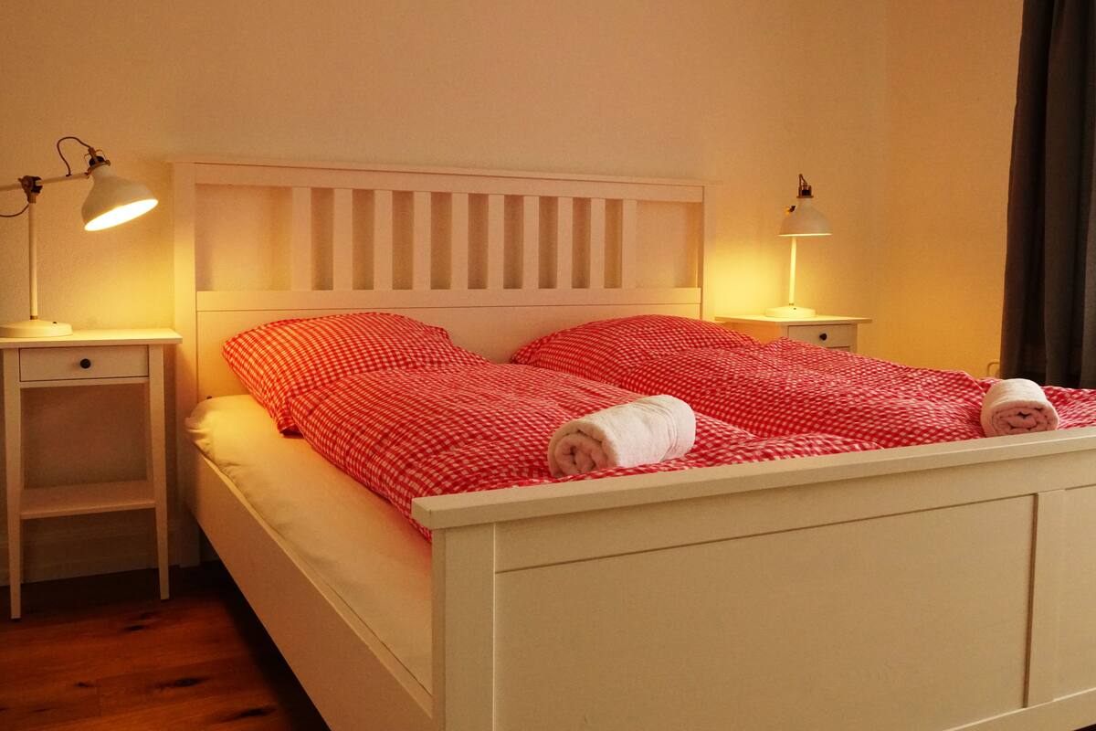 Uma cama com cabeceira branca e roupa de cama xadrez vermelha, duas luminárias de cabeceira e uma janela com cortinas.
