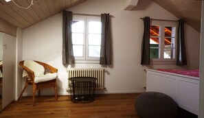 A room with a high ceiling, a wicker chair with a white cushion, a small round table, a radiator, and a window with curtains. - Adventure Guesthouse (Unterseen)