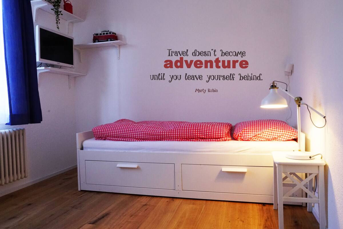 Um quarto pequeno e bem arrumado, com uma cama de casal, um abajur e uma frase motivacional na parede.