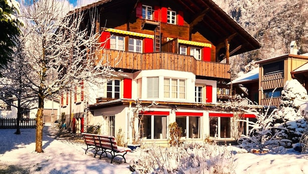 A two-story chalet with a wooden facade and red accents, surrounded by snow-covered ground and trees. - Adventure Guesthouse (Unterseen)