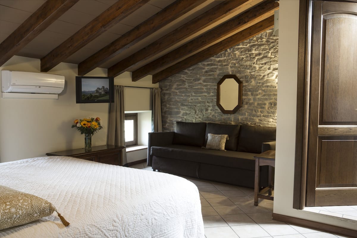 A bedroom with a large bed, a stone wall, a dark sofa, and a wooden ceiling.