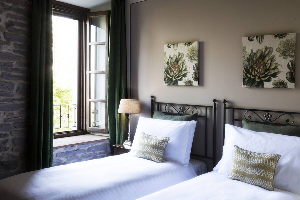 A bedroom with two beds, a window with curtains, a nightstand with a lamp, and floral wall art.