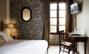 A bedroom with a stone wall, a bed, a chair, a desk, a lamp, a TV, and a window with curtains. - La locanda del Borgo (Langhirano)