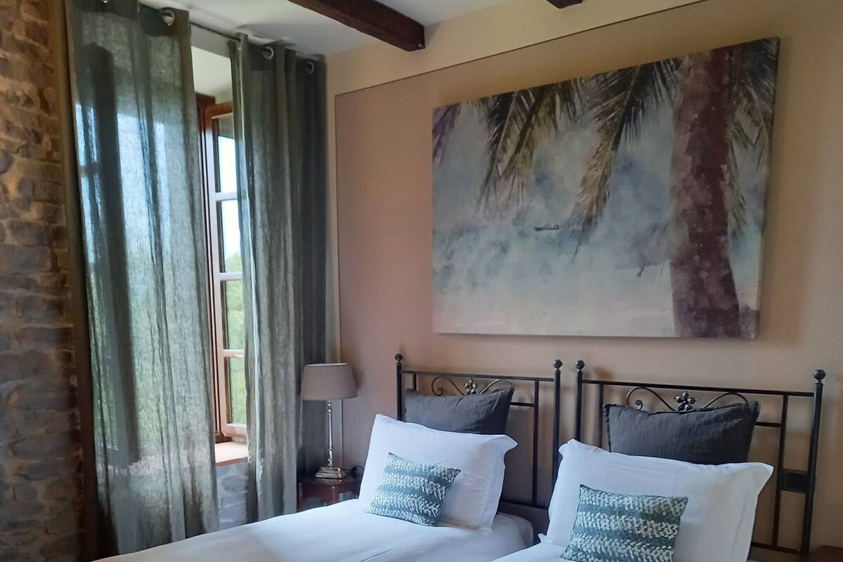 A double bed with white linens and two pillows, a nightstand with a lamp, a painting of palm trees, and a ceiling fan.
