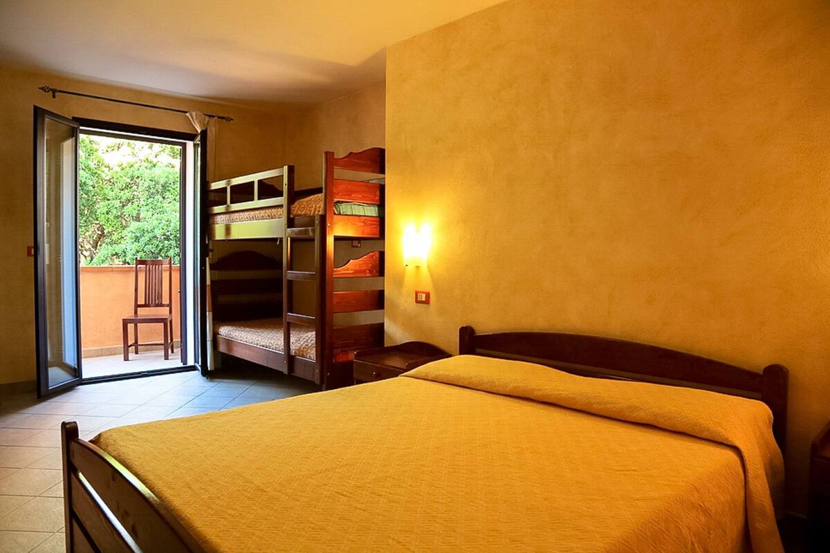 A room with a single bed, a wooden bunk bed, and a chair near a sliding glass door leading to a balcony.
