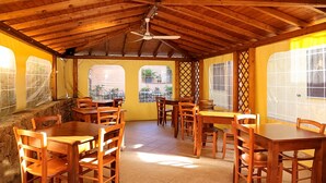 Un comedor interior con mesas y sillas de madera, un ventilador de techo y amplios ventanales con vistas al exterior.