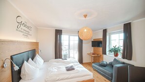 Un dormitorio moderno con cabecera de madera, una cama grande con ropa de cama blanca, un sofá gris, una mesita con una planta y un ventanal con cortinas.