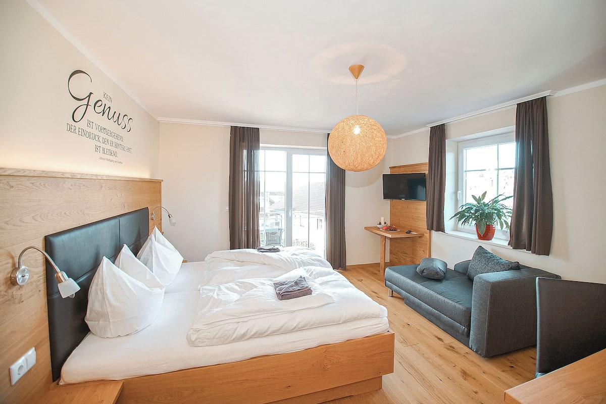 Un dormitorio moderno con cabecera de madera, una cama grande con ropa de cama blanca, un sofá gris, una mesita con una planta y un ventanal con cortinas.