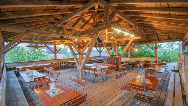 Una terraza de madera al aire libre con mesas y bancos, rodeada de árboles.