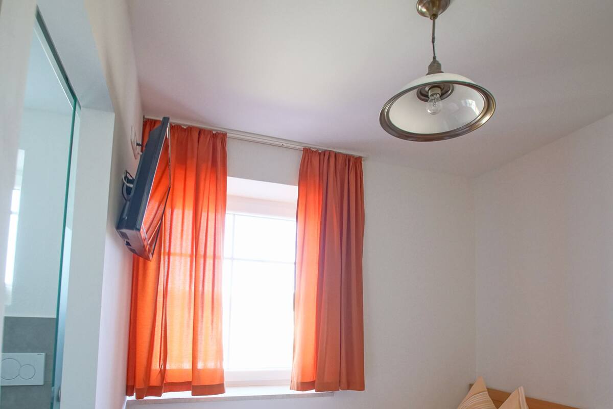 Una habitación pequeña con cama individual, suelo de madera, una ventana con cortinas naranjas y un ventilador de techo.