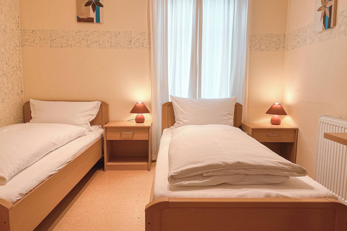 Habitación doble con dos camas individuales, mesitas de noche con lámparas, una ventana con cortinas y un radiador.