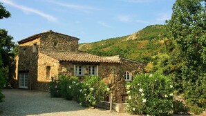 A stone house with a tiled roof, surrounded by gravel and flowering plants, set against a hillside. - Hameau des Claudins (Plan-de-la-Tour)