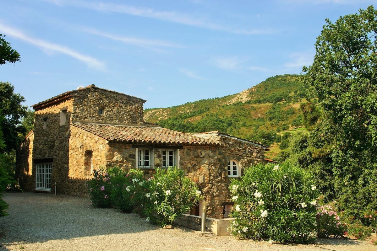 Une maison en pierre avec un toit de tuiles, entourée de gravier et de plantes en fleurs, située au pied d’une colline.