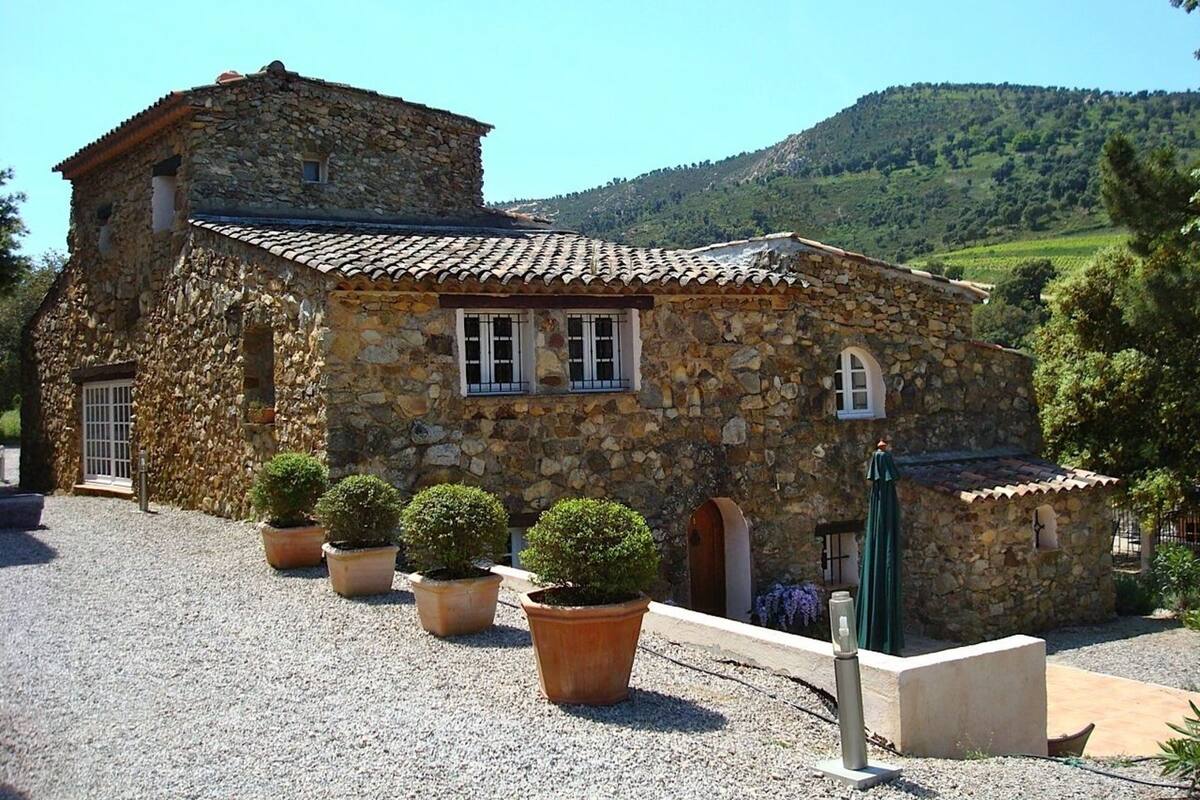 Un bâtiment en pierre avec un toit en tuiles, entouré de gravier, de plantes en pot et dominé par un paysage montagneux.