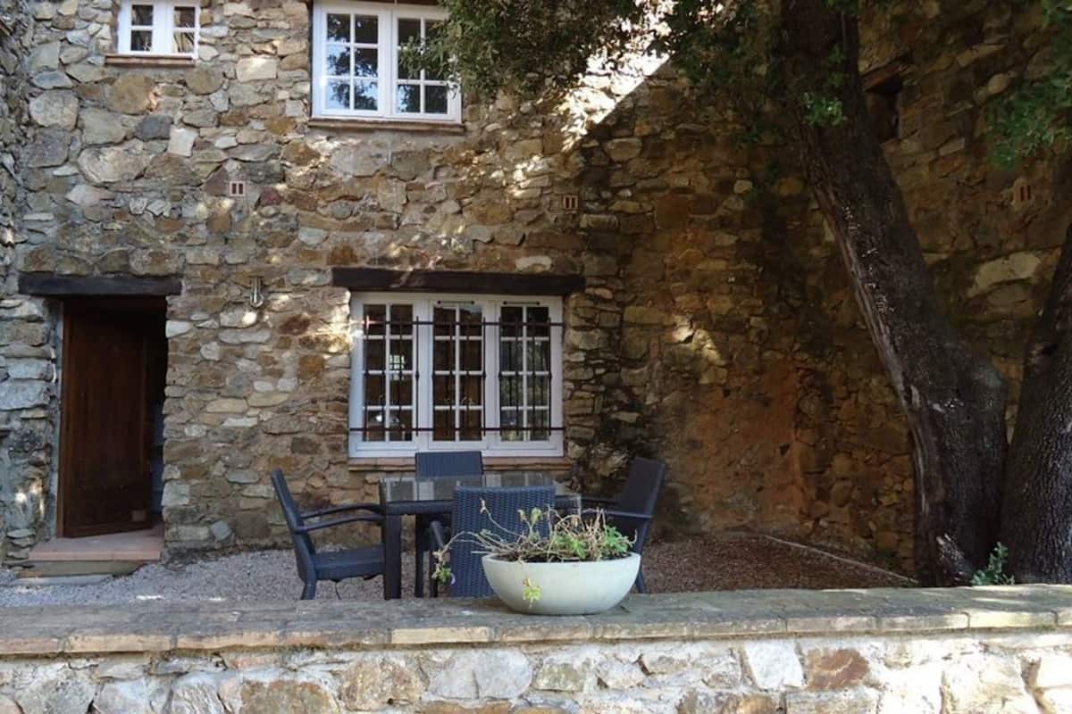 Un bâtiment en pierre avec une porte en bois, une fenêtre aux cadres blancs, une table avec des chaises et un pot de fleurs.