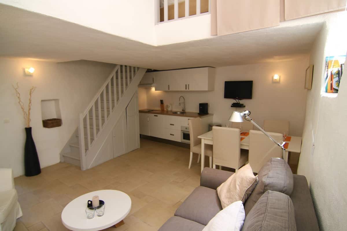 Un espace de vie compact comprenant un escalier menant à un niveau supérieur, une kitchenette, un coin repas avec une table et des chaises, et un coin salon avec un canapé et une petite table ronde.