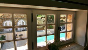 A view through a glass door with multiple windows, leading to an outdoor area with a stone wall, a pool, and outdoor furniture. - Hameau des Claudins (Plan-de-la-Tour)