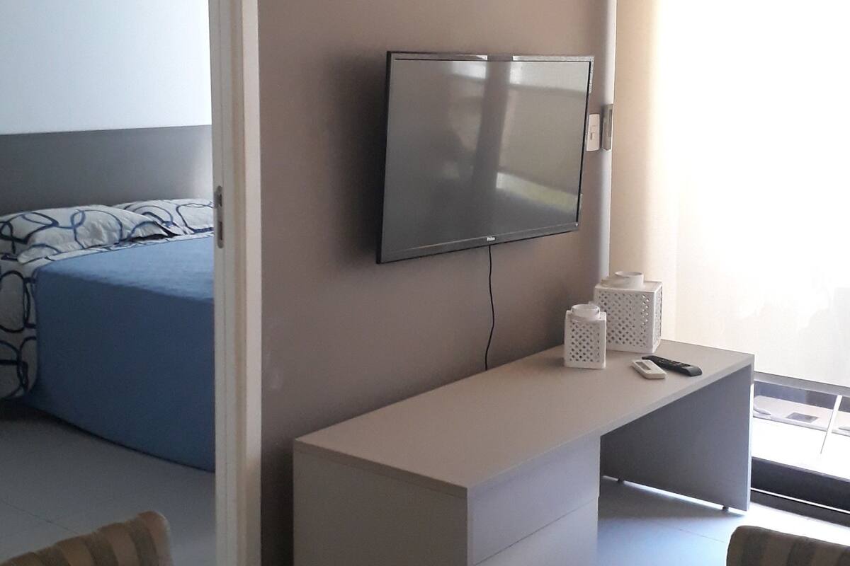 A modern hotel room with a flat-screen TV mounted on the wall, a small white side table, a glass dining table with chairs, and a bed with a blue and white patterned bedspread.