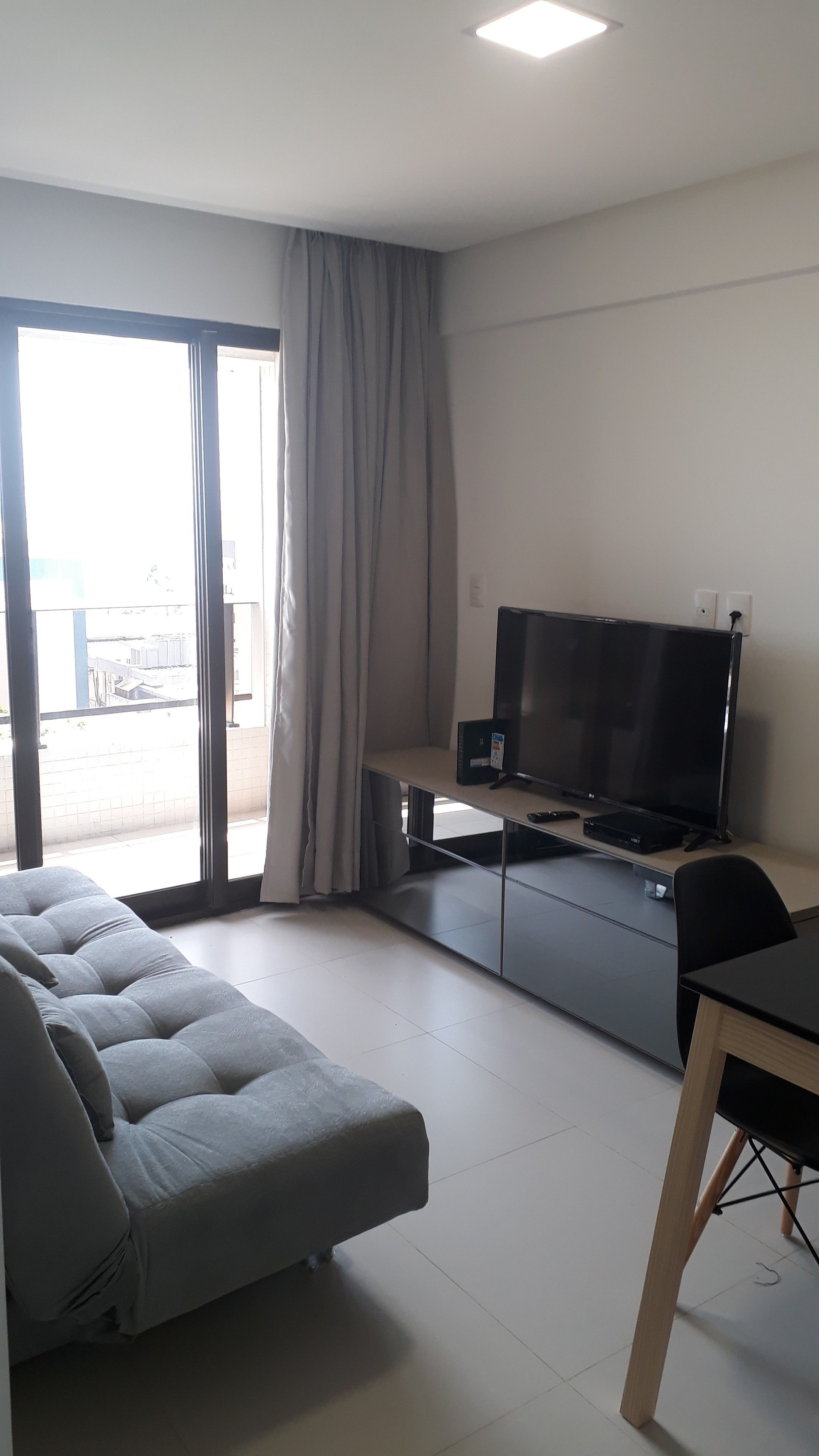 A modern living room with a grey sofa, a flat-screen TV, a dining table, and a sliding glass door leading to an outdoor area.