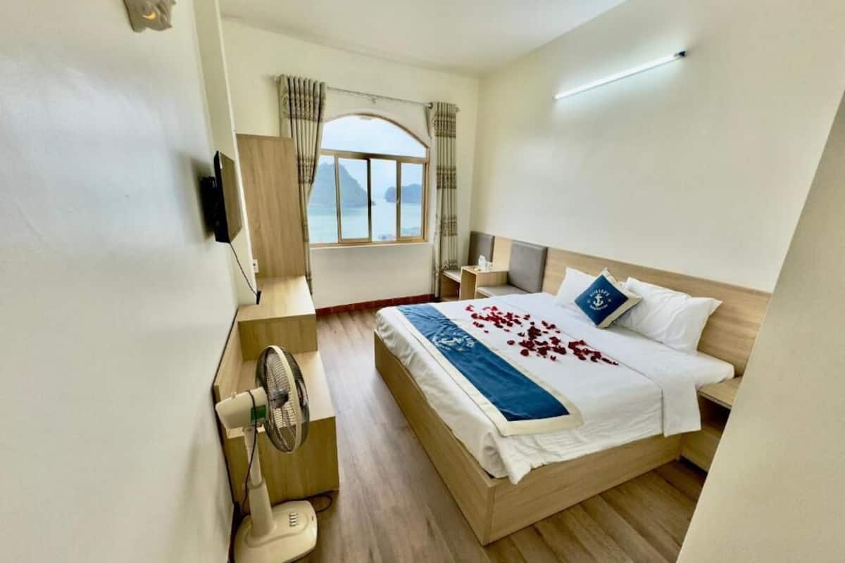 A hotel room with a bed, a bedside table, a fan, and a window with a view of the sea.