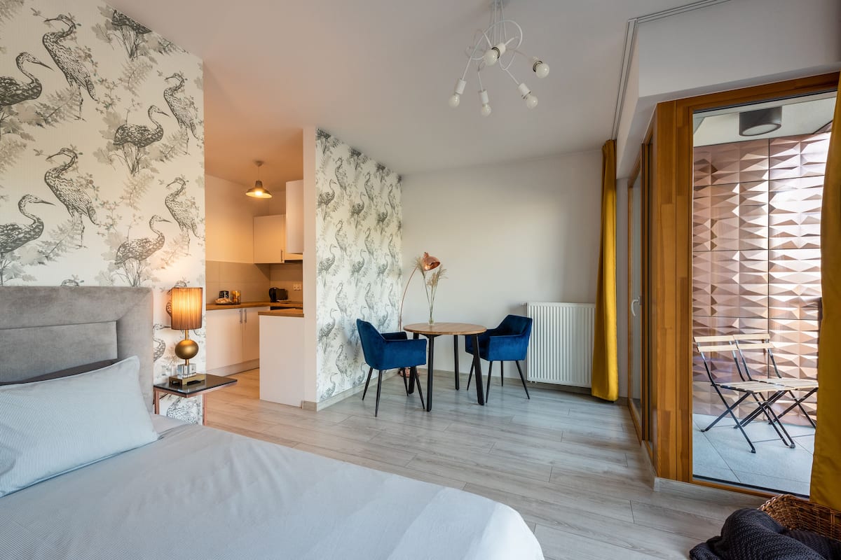 Une chambre d’hôtel moderne avec un lit, un coin repas avec des chaises bleues, une cuisine et une vue sur l’extérieur, visible à travers une porte vitrée.