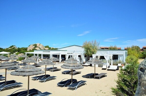 A beachside resort with sun loungers and thatched umbrellas, a white building with a green roof, and a rocky landscape in the background.