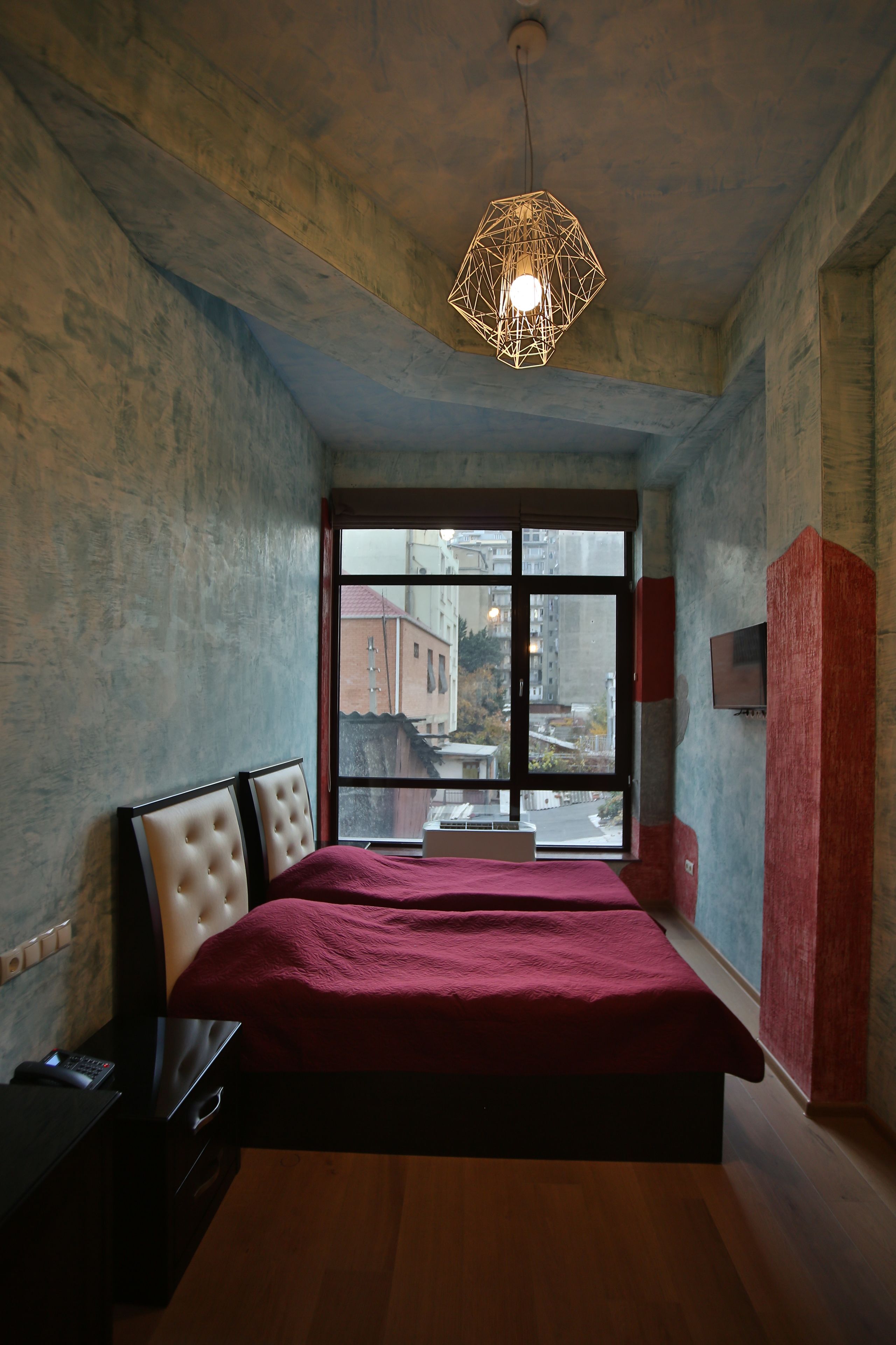 A bedroom with a large bed, a wooden floor, a window with a view of buildings, and a hanging light fixture.