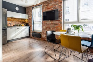 A modern kitchen with white cabinets, a brick wall, a flat-screen TV, and a dining area with yellow chairs.