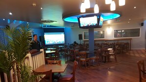 Un restaurante con mesas y sillas de madera, una zona de bar y una pantalla de televisión mostrando un programa.