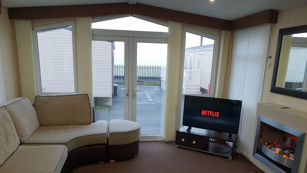Una sala de estar con un sofá color beige, un televisor de pantalla plana mostrando 'NETFLIX', una chimenea y vistas a un balcón con una valla blanca.