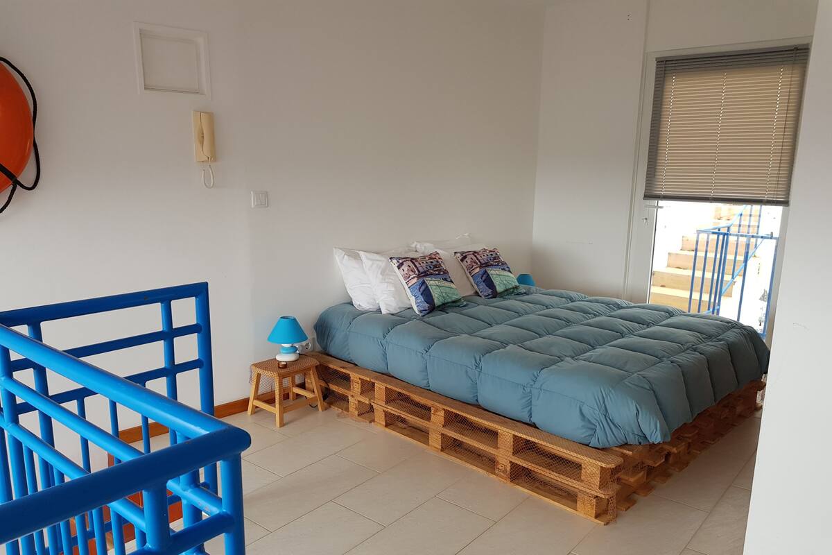 A bedroom with a blue bed, a small wooden nightstand, a blue chair, and a window with blinds.