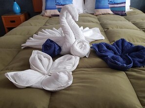 Un lit recouvert d’une couette verte, des serviettes blanches et bleues disposées de manière décorative, et une petite table de chevet en bois sur laquelle se trouve un vase bleu.
