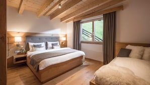 Ein Schlafzimmer mit Holzbalkendecke, einem großen Bett mit grauem Kopfteil, einem Nachttisch mit Lampe und einem Fenster mit Blick auf einen Balkon.