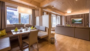 Ein gemütlicher, gut beleuchteter Raum mit Holzbalkendecke, einem Essbereich mit Tisch und Stühlen, einer Couch und einem großen Fenster mit Blick ins Freie.