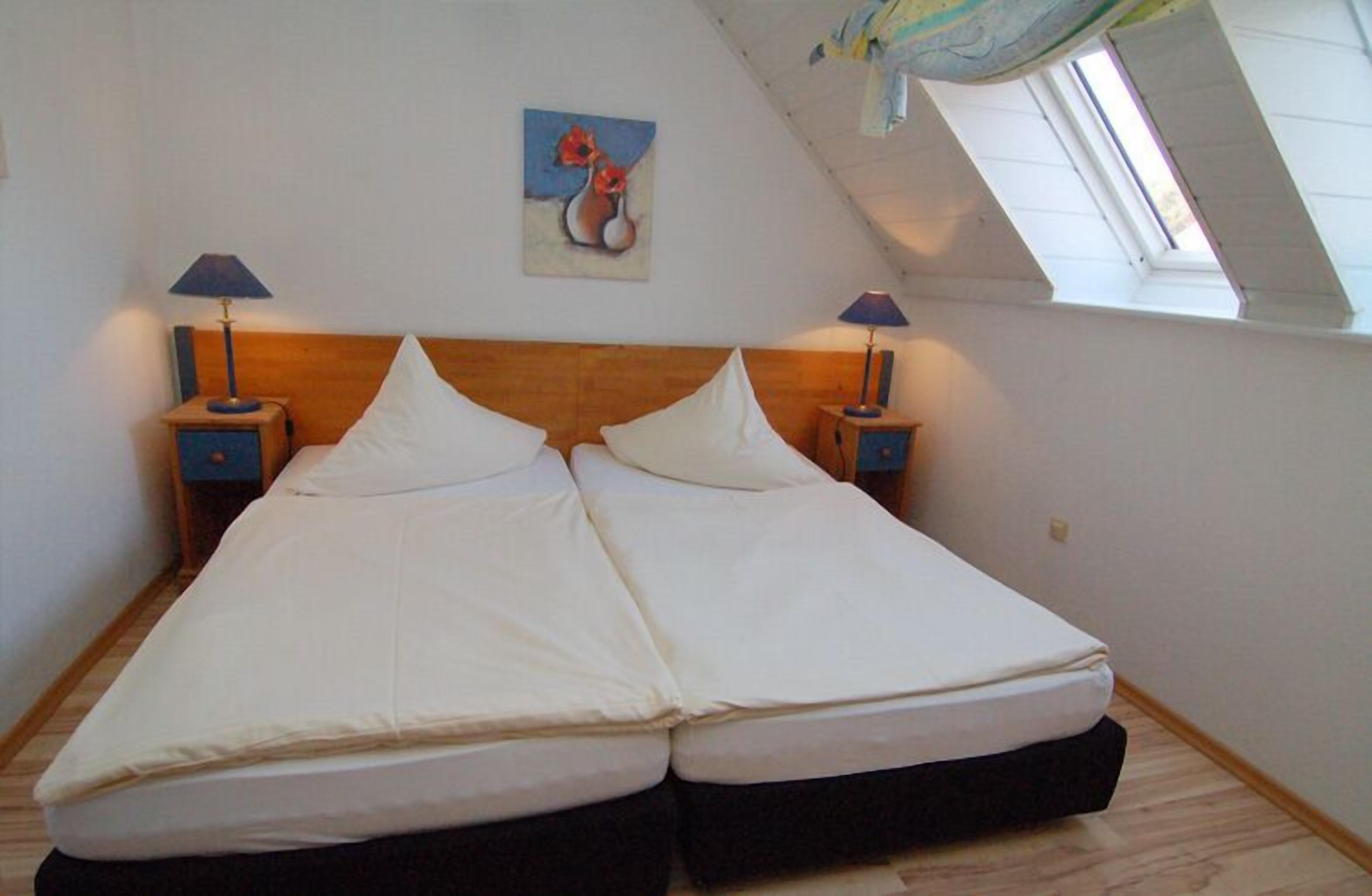 A double bed in a room with a sloped ceiling, two bedside tables with lamps, and a painting on the wall.
